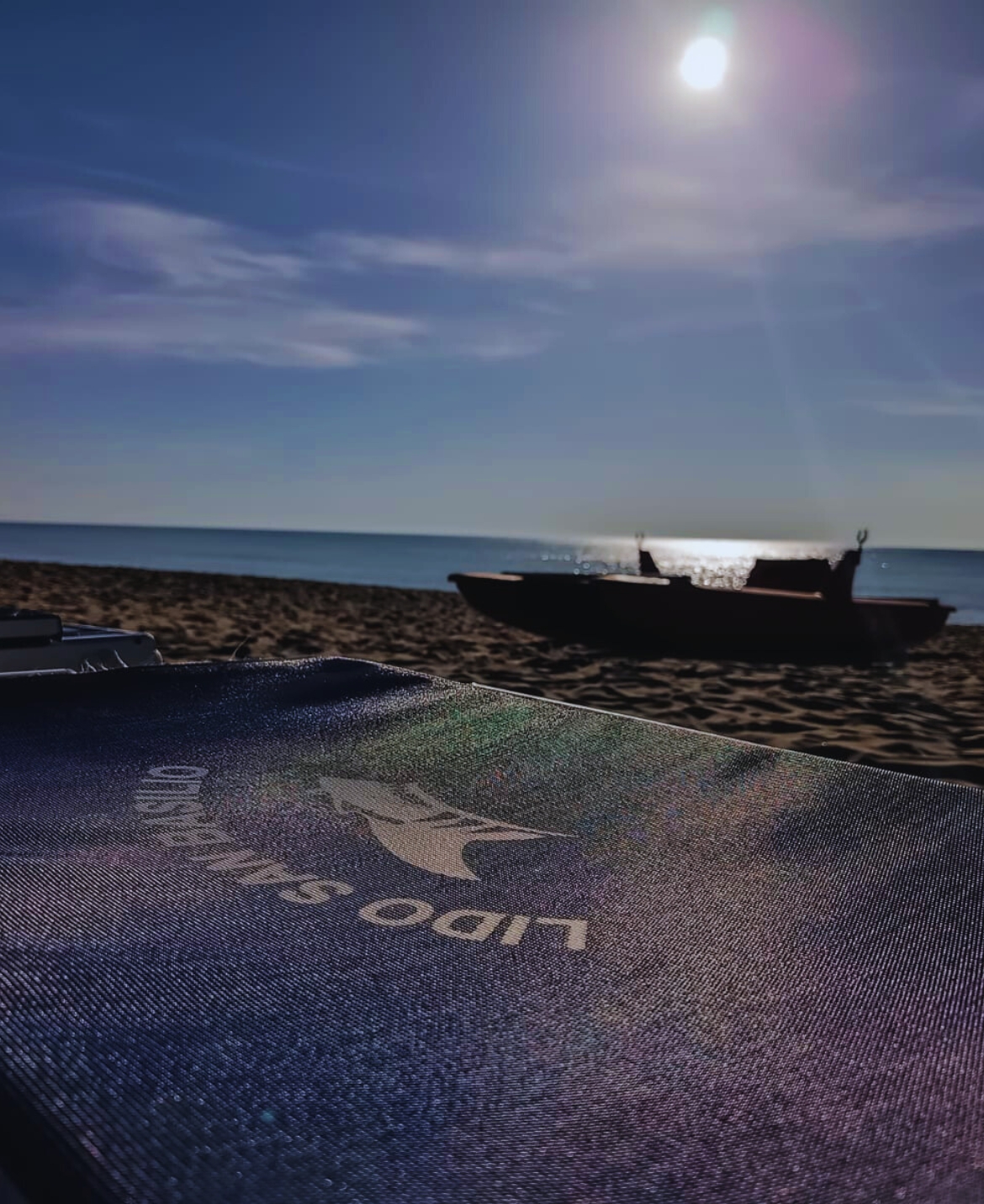 Ombrellone con logo Lido San Basilio in primo piano e barca sulla spiaggia al tramonto